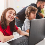 young girl sitting in classroom with her computer next to a young boy and teacher, smiling with her laptop using innovative assessment tools.