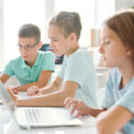 Three young school kids in casual wear sitting in computer classroom using laptops.