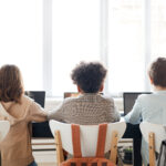 View from behind of three children sitting in a row in front of their computers.